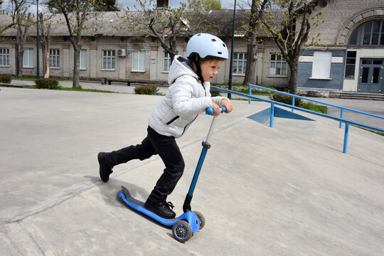 Young boy riding a blue scooter at a skatepark wearing a helmet and