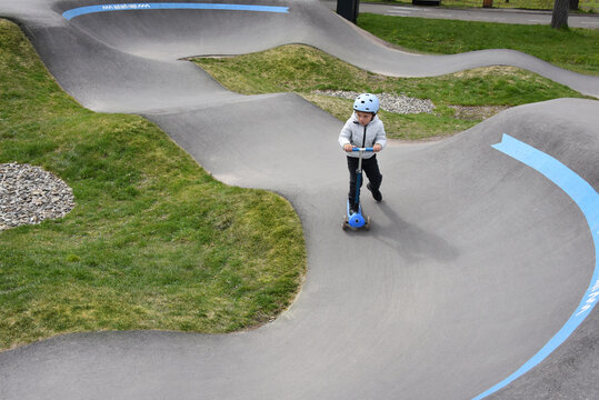 A young child rides a blue scooter along a winding pump track in a