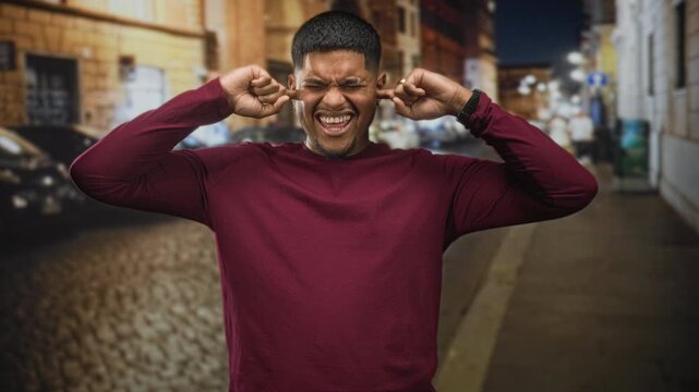 Hispanic man plugs ears with fingers on a night street lined with cobblestone and parked cars; frustration sensory overload.