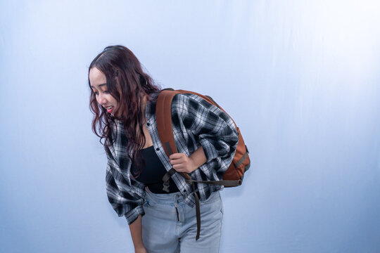 Tired Asian female student with heavy backpack leaning forward on plain background
