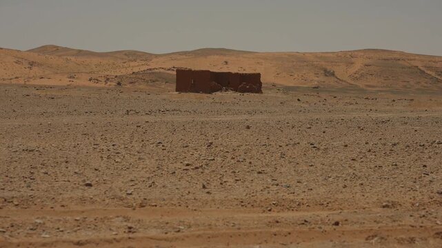Lone ruined mud brick structure sits isolated on rocky desert plain with rolling sand hills behind