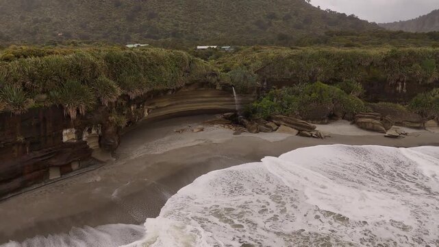 Drone flying backwards from a coastal waterfall on a beach in Truman Track, West Coast of South Island, New Zealand