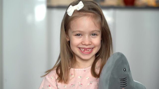 Portrait of little happy toothless girl with toy shark looks at camera and laughs. Childrens oral hygiene. Baby milk teeth change. Health care