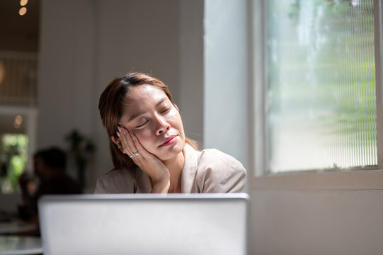 Asian woman closing eyes resting head on hand under sunlight sitting with laptop beside cafe window.