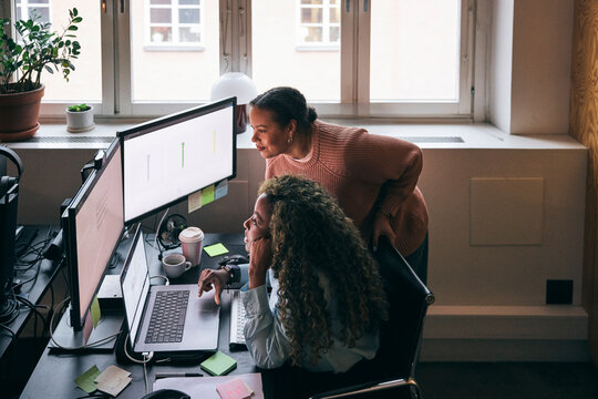 High angle view of multiracial female tech professionals comparing responsive design preview on dual monitor in startup