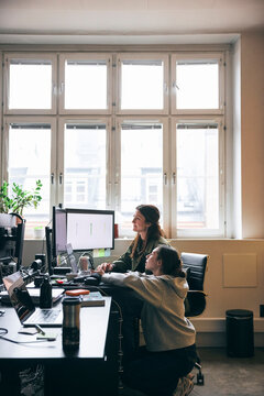 Female tech professional crouching near coworker while discussing data previews on devices at office