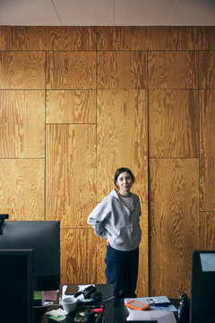 Portrait of female tech entrepreneur standing with hands in pockets against brown wall in startup office