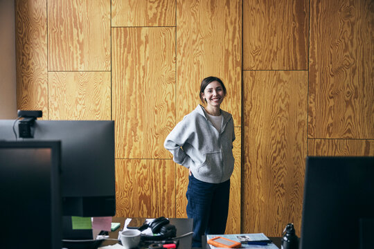 Portrait of smiling female tech entrepreneur standing with hands in pockets against brown wall in startup office