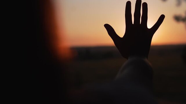 Person raises hand toward sunset creating silhouette at horizon. Human reaches palm against golden sun at dusk. Hand blocks sunlight. Person shows gesture at sunset. Silhouette reaches toward sun.