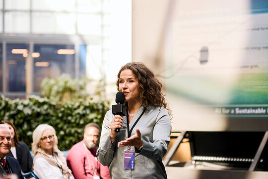 Mature female speaker explaining presentation points while speaking into microphone during summit in congress hall