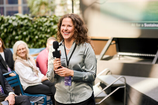 Smiling female presenter holding microphone while standing for speech during seminar in business convention hall