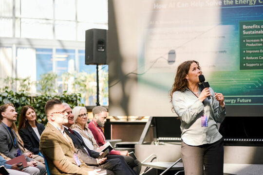 Mature female presenter discussing presentation points with audience while speaking into microphone during summit in con