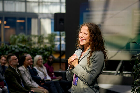 Smiling mature female presenter communicating with audience at seminar in convention center