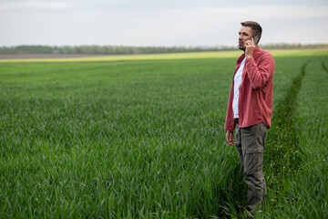 Farmer standing in a green wheat field and talking on mobile phone during agricultural work. Agronomist communicating while monitoring healthy wheat crop growth in farmland.  © Тетяна Шустик