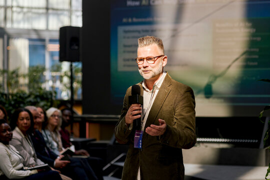Mature male keynote speaker using microphone while communicating with audience during summit in congress hall