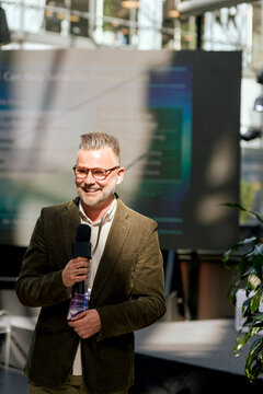 Smiling mature male keynote speaker using microphone while communicating during summit in congress hall