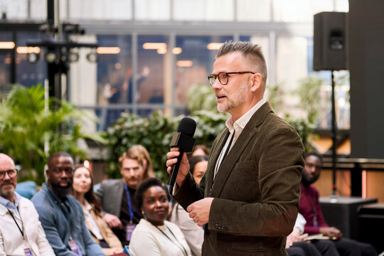 Male speaker wearing eyeglasses holding microphone while addressing business audience during seminar in convention hall
