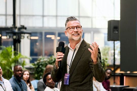 Smiling male presenter holding wireless microphone delivering keynote speech during business seminar in congress center