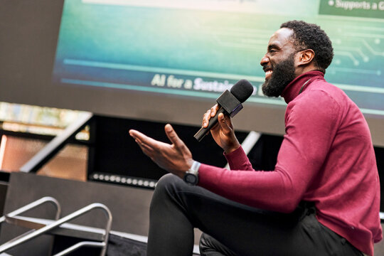 Happy male presenter communicating through microphone while sitting on platform during presentation in convention center