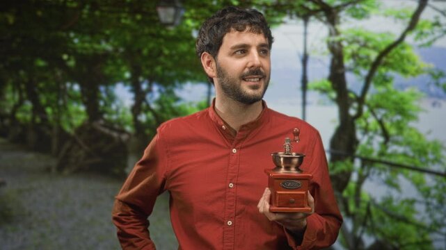 Young hispanic man with beard holds a vintage coffee grinder in right hand on a forest path beside a lamp post and metal railing; contentment.