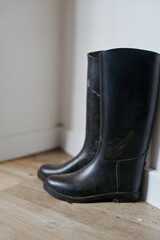 Fototapeta premium A pair of black rubber boots rests beside a white wall on a wooden floor, suggesting ready outdoor wear in a simple home scene with quiet, everyday routine and anticipation