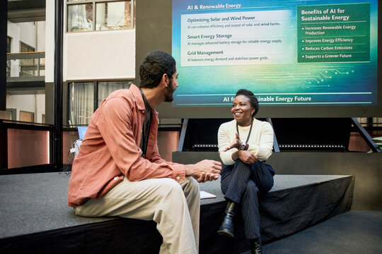 Smiling businesswoman talking with male business executive while sitting on platform near projection screen in conventio