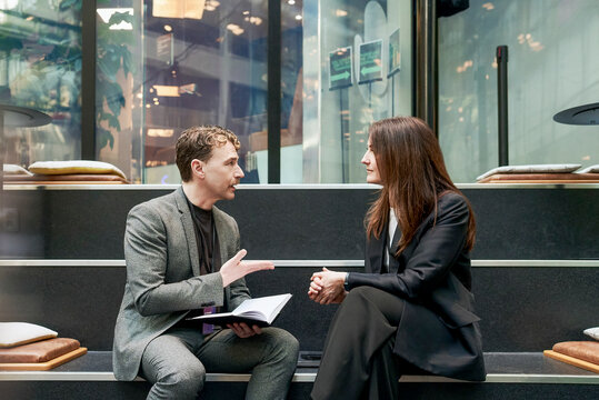 Mature businessman discussing ideas with female expert while sitting on step in networking event at convention center