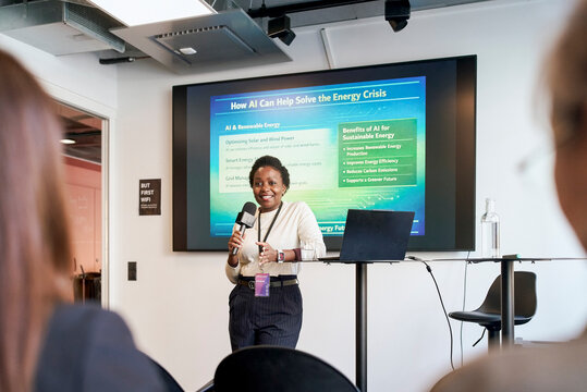 Smiling female business professional holding microphone while addressing audience and giving presentation at seminar in