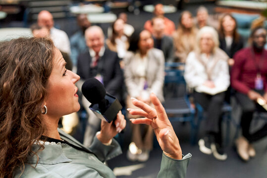 Headshot of female keynote speaker holding microphone while talking at business summit in congress hall