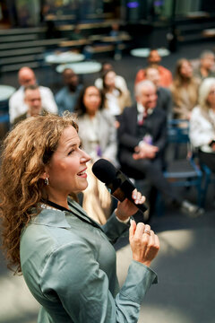 High angle view of smiling female speaker speaking into microphone during business summit in convention hall