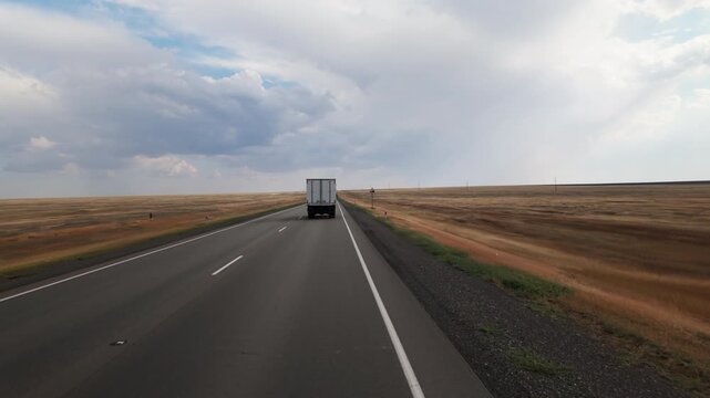 Truck with white semi-trailer van is driving along road between fields in steppes