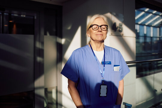 Senior female nurse day dreaming while standing with eyes closed in medical building