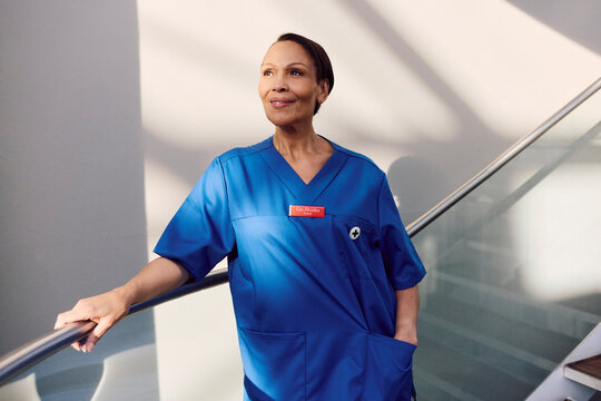 Smiling short haired female medical expert looking away while standing with hand in pocket near railing of hospital