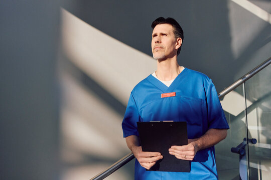Thoughtful mature male medical expert holding note pad standing near railing at hospital