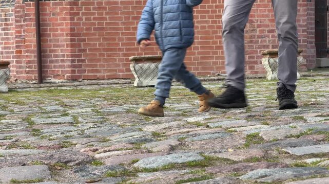 low angle view of adult legs in grey jeans and black boots walking hand in hand with child in blue wet pants and beige shoes on red brick street wet cobblestone rainy day urban scene no faces visible