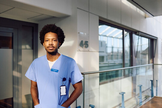 Portrait of male medical professional standing with hands in pockets at hospital corridor