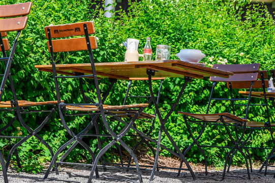 Schwangau, Germany &mdash; June 04 2026: Detail view of an outdoor wooden cafe table and folding chairs in a beer garden with green bushes in the background near Hohenschwangau.