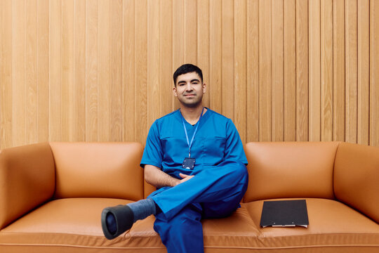 Portrait of young male healthcare professional sitting on sofa against wooden wall in hospital lobby