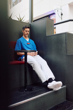 Full length of mature male healthcare worker spending break time using smart phone while sitting in hospital cafeteria