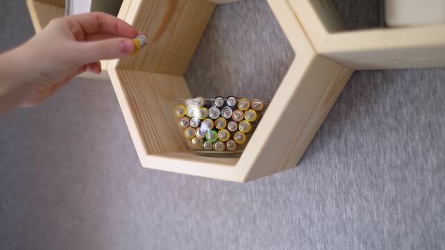 A young woman places a glass bowl filled with used batteries onto a wooden shelf for future disposal. This process demonstrates conscious household waste sorting and care for the environment.