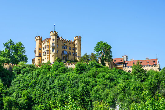 Schwangau, Germany &mdash; June 04 2026: Low angle view of the picturesque Hohenschwangau Castle rising above a lush green forest under a clear blue sky.