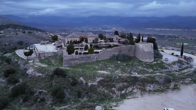 Aerial tracking shot circling the stone fortification walls, round defensive tower, and central restaurant building of Lekuresi Castle overlooking Sarande city at dusk, Albania