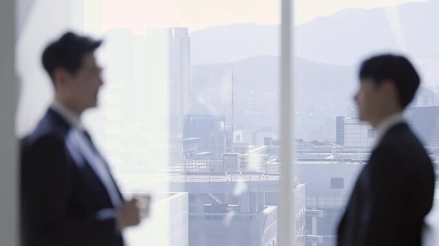 Blurred silhouettes of two businessmen in suits by a large office window, overlooking a modern cityscape with distant mountains, suggesting a corporate environment or business discussion.