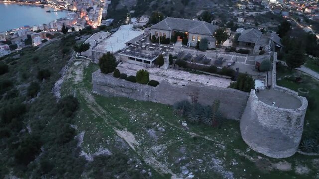 Aerial tracking shot circling a prominent stone round tower and the fortress walls of Lekuresi Castle complex overlooking Sarande bay and city lights at dusk in Albania