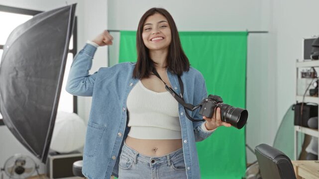 Woman holding camera and flexing arm in studio with green screen backdrop and softbox lighting; confidence empowerment.