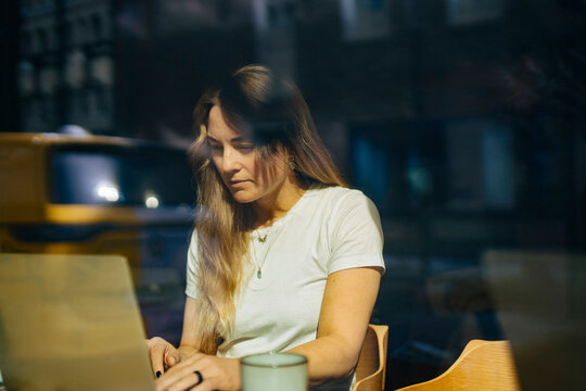 Focused businesswoman with long hair working on laptop behind glass window at coworking space