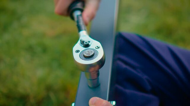 Hands applying torque with socket wrench fixing nut on metal support bar outdoors. Structural assembly workflow with durable materials and tool control in daylight. Industrial maintenance concept with