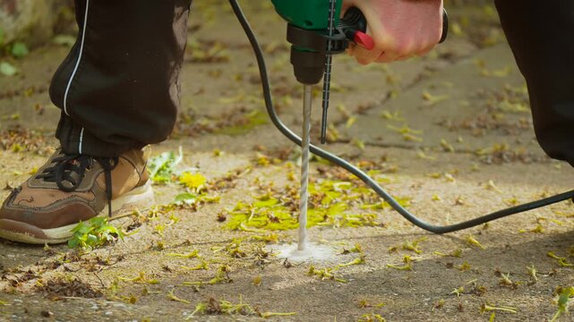 Rotary hammer drilling concrete ground creating dust cloud around bit close perspective. DIY builder operating corded drill for fixing anchors in cement walkway outdoor setting. Maintenance repair