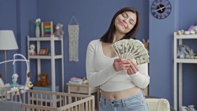 Woman fanning dollar bills with both hands, bare navel visible, smiling in a baby nursery with crib and mobile; financial relief joy.