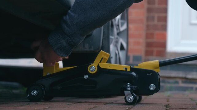 Positioning hydraulic floor jack with wooden spacer beneath vehicle for car lifting preparation. Automotive maintenance concept with lifting equipment, wheel area access on driveway. DIY vehicle
